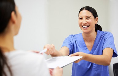 Patient handing completed forms to woman in blue scrubs