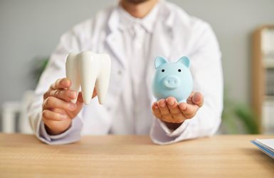 Dentist in white jacket holding model tooth in one hand and blue piggy bank in other