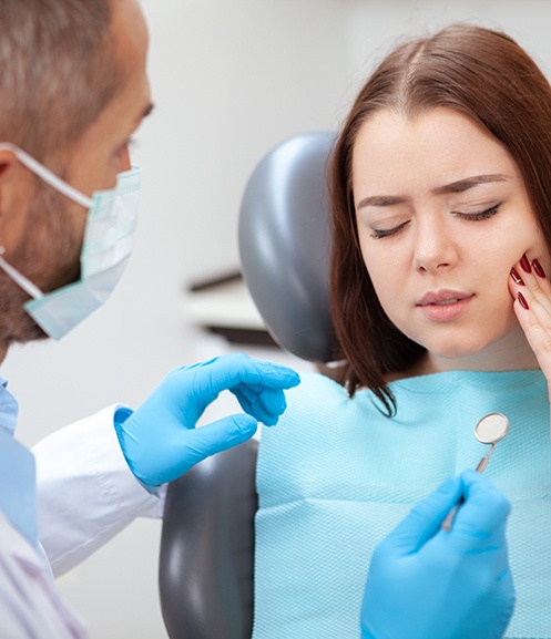 Woman in dental chair holding hand to chin with eyes closed in pain